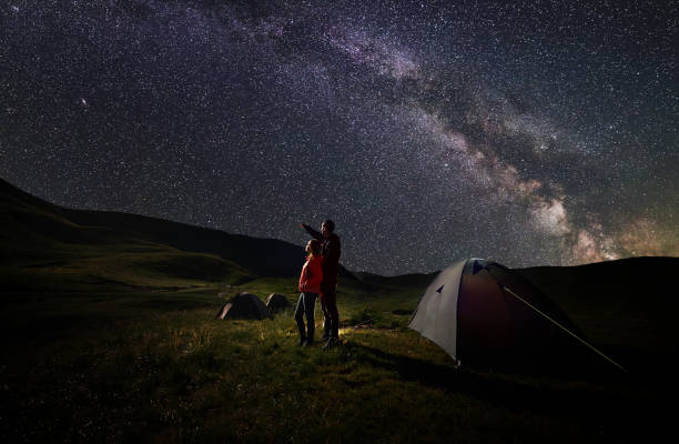 Photo of Guy and girl stand near tents under starry sky against backdrop of mountains covered by greenery and lake