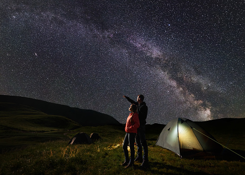 Male shows female up on the night starry sky at Milky way. Happy pair backpackers standing near illuminated tent at camping, against the backdrop of the mountains covered by greenery and the lake.