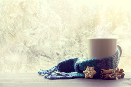 Cinnamon, gingerbread and white mug wrapped in a scarf, stands on a table on the background of a frozen window