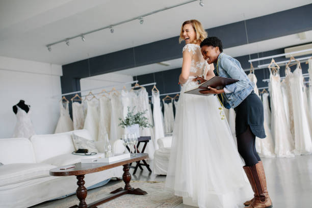 Female trying on wedding gown with women assistant in shop Female trying on wedding gown with women assistant in bridal wear shop. Smiling woman wearing her bridal dress with wedding dress designer in bridal fashion boutique. bridal shop photos stock pictures, royalty-free photos & images