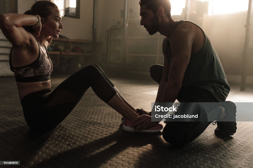Male coach assisting woman in doing sit-ups at gym Male coach assisting woman in doing sit-ups at gym. Woman working out on her abs with help from male personal trainer. Fitness Instructor Stock Photo