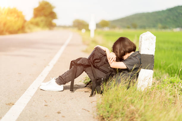 Young asian woman short hair and wearing sunglasses sit with backpack hitchhiking along a road wait for help in countryside Thailand