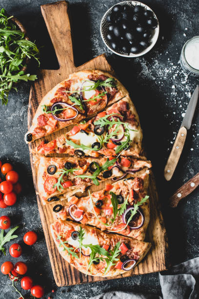 istock Flatbread pizza garnished with fresh arugula on wooden pizza board, top view 941400068