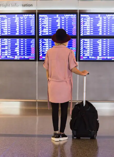 Photo of Young pretty woman in international airport looking at the flight information board with carry on luggage