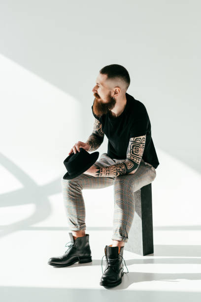 stylish tattooed man sitting and holding hat on white