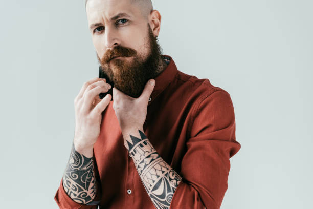 istock tattooed man combing beard and looking at camera isolated on white 941420496
