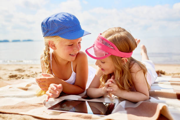 Cute girls Curious little friendly girls with touchpad lying on sandy beach on hot day and watching videos girls laying on beach stock pictures, royalty-free photos & images