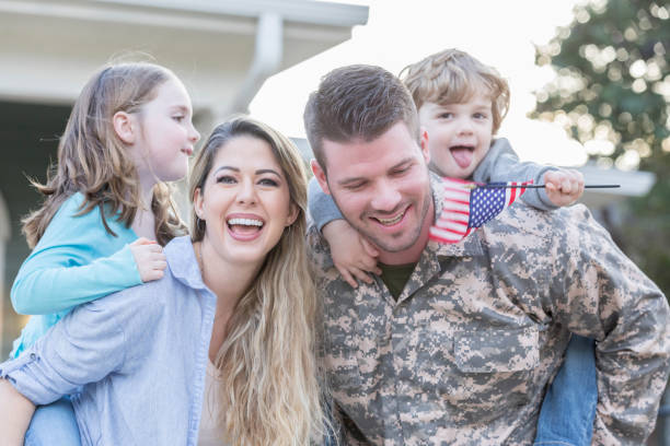 Happy family reunion Excited family is happy to have their husband and father home from overseas deployment. The couple are carrying their children on their backs. A toddler boy is holding a US flag. proud army mom stock pictures, royalty-free photos & images