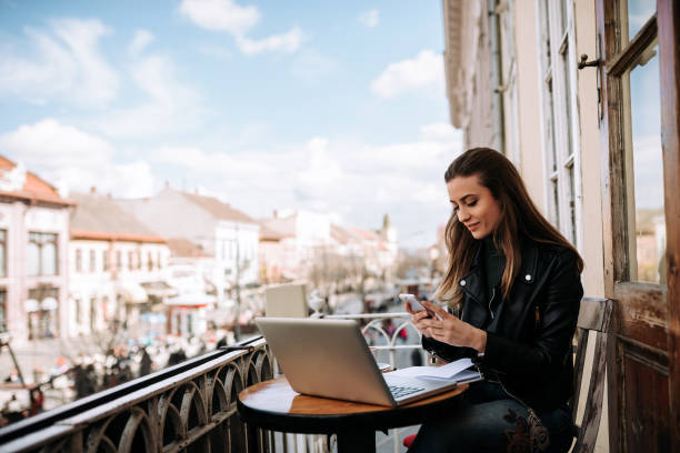 istock Young woman working on a balcony. Using smartphone and laptop computer outdoors. 941440514