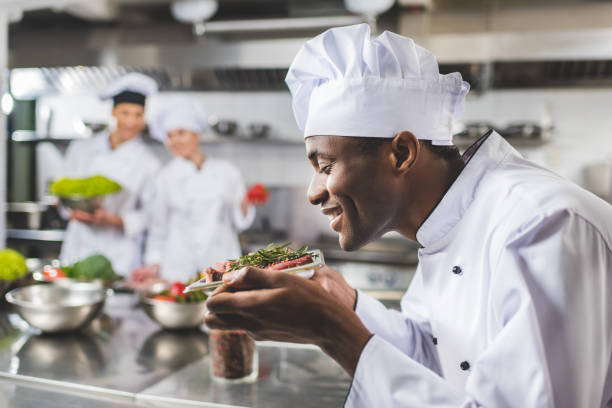 souriant chef afro-américain renifler la viande crue aux herbes à la cuisine du restaurant - chef restaurant cooking african descent photos et images de collection
