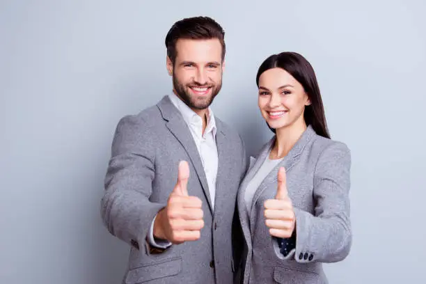 Photo of Two confident smiling businesspeople in formalwear showing thumbs-up on gray background