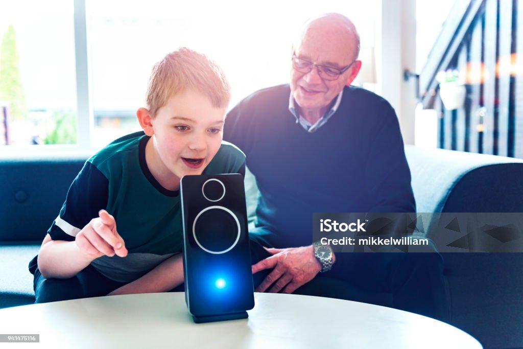 Boy speaks to digital assistant while his grandfather look at him A boy sits in a sofa with his grandfather and they are bonding with help of consumer electronics. In front of them on a table stand a digital assistant, which is an intelligent loudspeaker with built-in microphone. The digital assistant can help answer questions and enable the family to control internet of things such as lightbulbs and door locks with voice commands. Voice Stock Photo