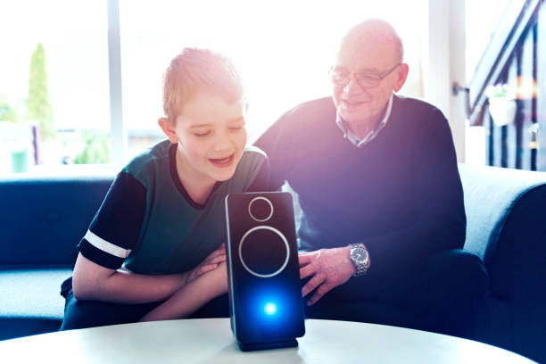 Digital assistant uses voice recognition to help family control internet of things at home A boy sits in a sofa with his grandfather and they are bonding with help of consumer electronics. In front of them on a table stand a digital assistant, which is an intelligent loudspeaker with built-in microphone. The digital assistant can help answer questions and enable the family to control internet of things such as lightbulbs and door locks with voice commands. smart home family stock pictures, royalty-free photos & images