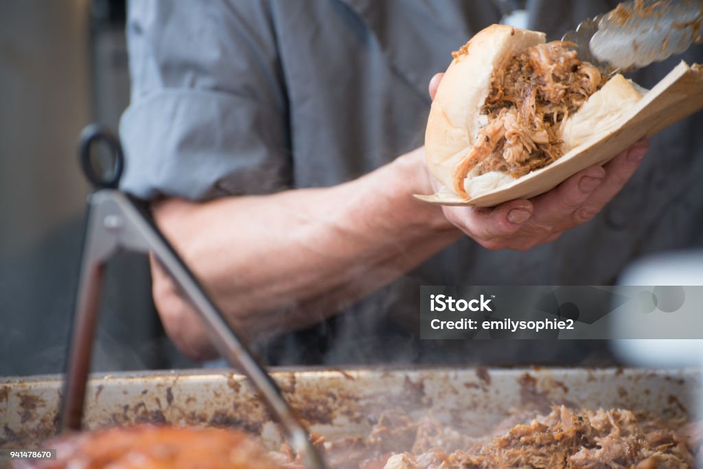 man preparing sandwich with fresh hot meat from serving tray Man in grey T shirt serving pulled pork street food holding sandwich with rind and serving tongs in the foreground Pig Stock Photo