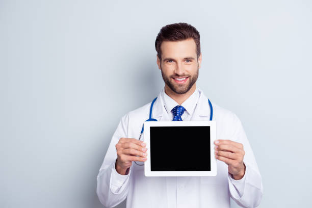 intelligent jeune médecin souriant en blouse blanche, ce qui démontre l’écran de sa tablette numérique sur fond gris - doctor digital display digital tablet healthcare and medicine photos et images de collection