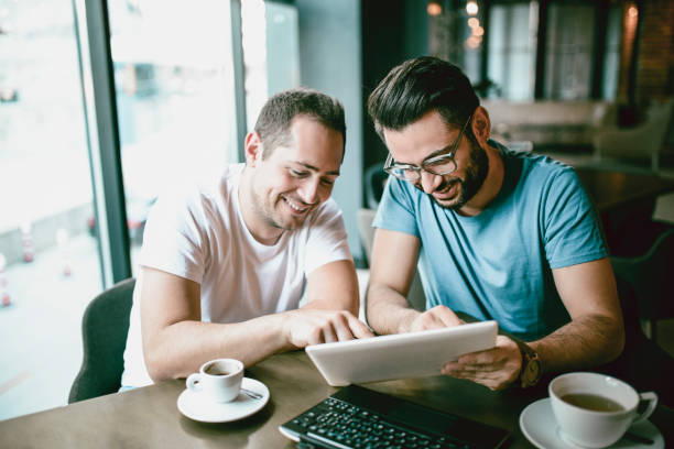 Handsome Smiling Freelancer Showing Finished Project on Digital Tablet to a Colleague in Coffee Restaurant