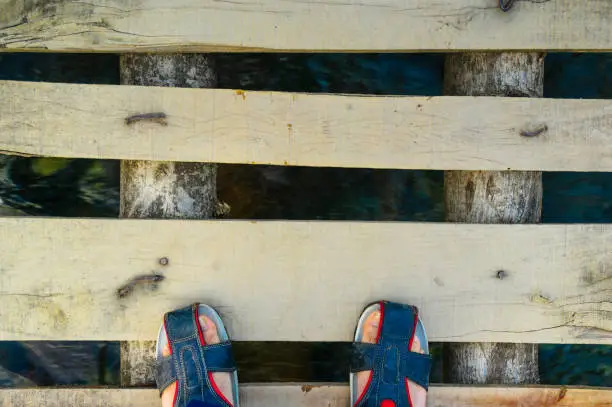 Feet in fashionable sandles standing on a wooden plank bridge with water flowing under it. Shows the bridges in rural areas or in hike or trails