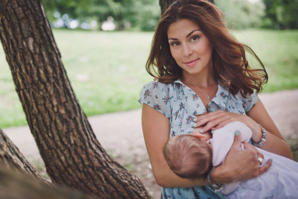 A beautiful smiling young brunette woman with long hair is breastfeeding a pretty baby. They are in the green summer forest. stock photo