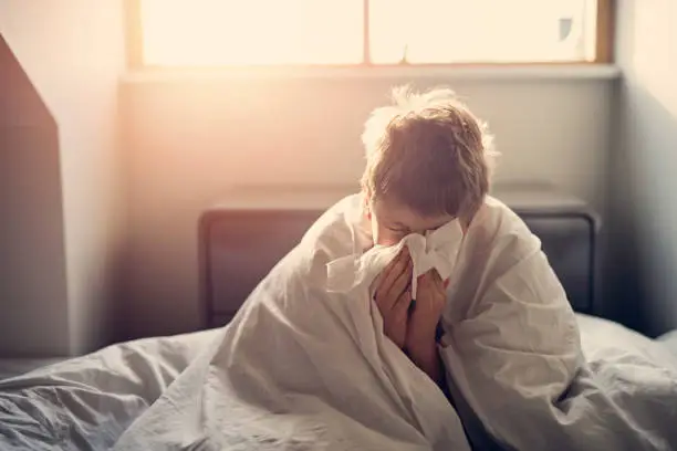 Photo of Sick little boy lying in bed and blowing nose