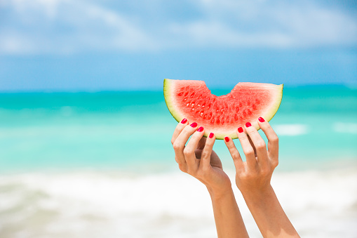 Woman holding slice of watermelon on the beach.