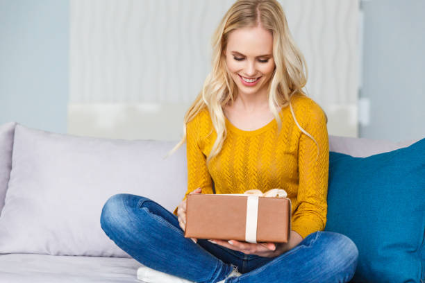 istock beautiful smiling young woman holding gift box while sitting on sofa at home 941686672