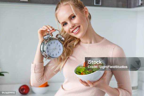 Fröhliche Junge Frau Holding Schüssel Mit Frischen Gesunden Salat Und Wecker Stockfoto und mehr Bilder von Essen - Mund benutzen