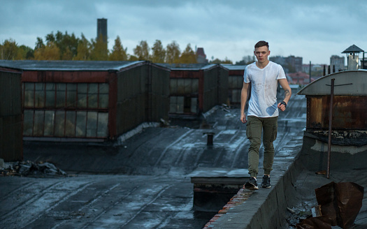 The young man is walking on a roof of a old industrial building. He is looking thoughtfully at the camera. The young man dressed only in a T-shirt and trousers at windy and cold autumn evening