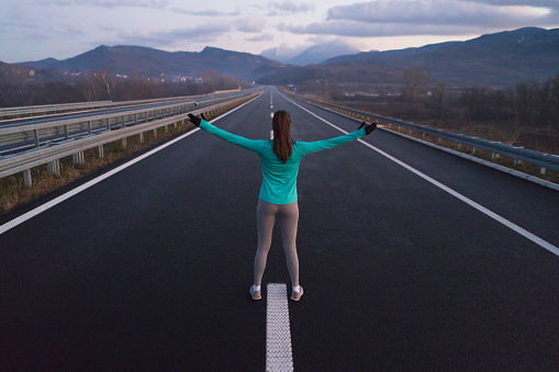 Rear view of a young female athlete standing on the highway with her arms outstretched.