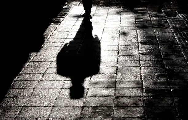 Blurry shadow and silhouette of a man standing in the night on wet city street sidewalk with water reflection in black and white