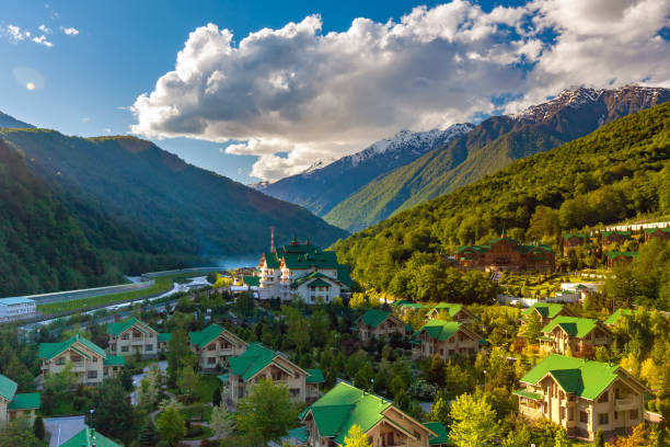 Ski Resort at Caucasus Mountains, Krasnaya Polyana, Sochi, Russia. Modern mountain cottages with green roofs against the backdrop of a mountain valley covered with a green spring sun forest. sochi stock pictures, royalty-free photos & images