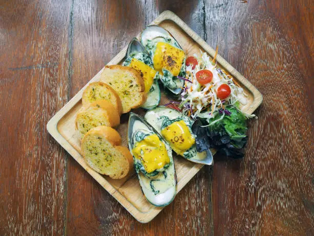 Photo of A delicious baked mussels with cheese and spinach served with butter bread and fresh salad on wooden tray.