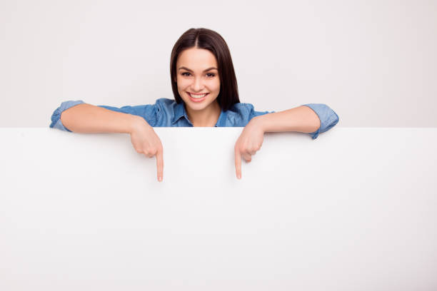 istock Cheerful cute girl is standing behind the white blank banner and pointing down at a copyspace 941737680