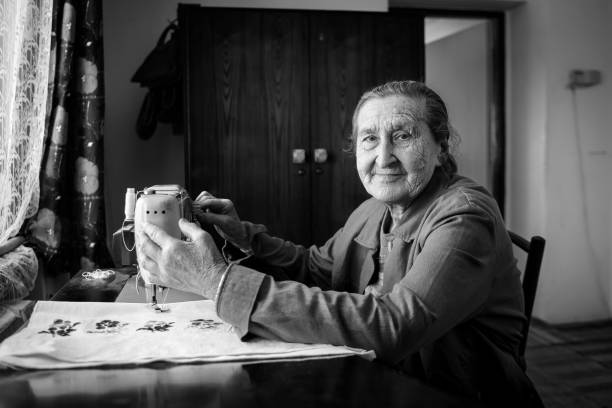 Cute 80 plus year old senior woman using vintage sewing machine. Black and white image of adorable elderly woman sewing clothes in her old family home.
