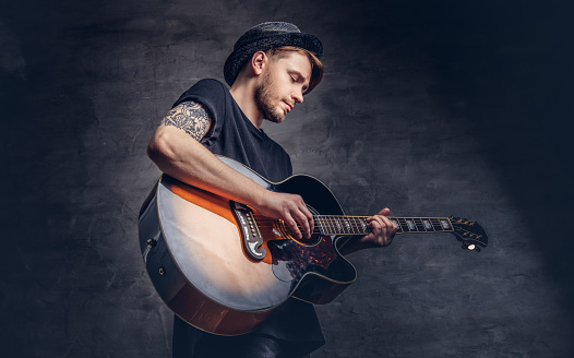 An attractive tattooed guitarist in hat playing on acoustic guitar, isolated on a dark background.