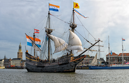 Old VOC sailing ship Halve Maen at the river IJssel during the 2018 Sail Kampen event in the Hanseatic league city of Kampen in Overijssel, The Netherlands. The Halve Maen was a trading ship of the Dutch East India Company (Dutch: Verenigde Oost-Indische Compagnie or VOC) and sailed into what is now New York Harbor in September 1609 during a search for a western passage to China. People on board are looking at the view and a crowd on the quay is watching the ships.