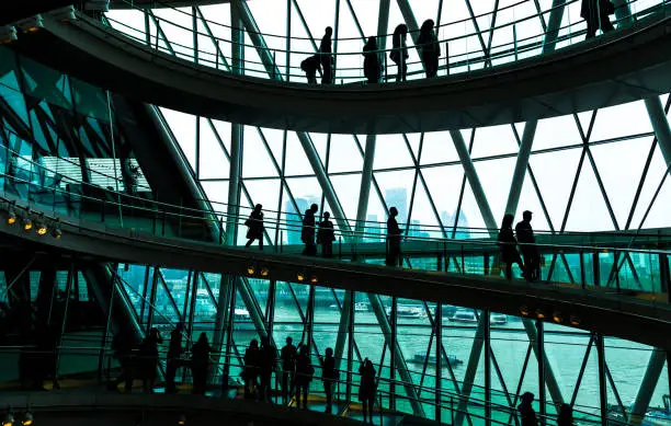 Wide angle color image depicting ultra modern contemporary interior architecture in City Hall (a public building in London that is open to the public) in London, UK. Crowds of people in silhouette and unrecognizable are walking down the modern spiral staircase. In the background we can see some of the skyscrapers that dominate the skyline of London. Lots of room for copy space.