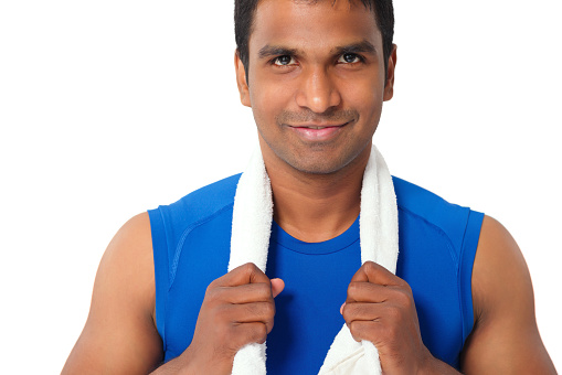 Head and shoulders portrait of smiling Indian sportsman with towel over his neck standing against white background and looking at camera