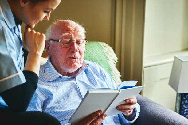 Mature man reading a book with home caregiver Mature man reading an interesting book with female caregiver at home hospice photos stock pictures, royalty-free photos & images