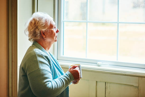 istock Pensive senior woman standing by a window with coffee 941789740