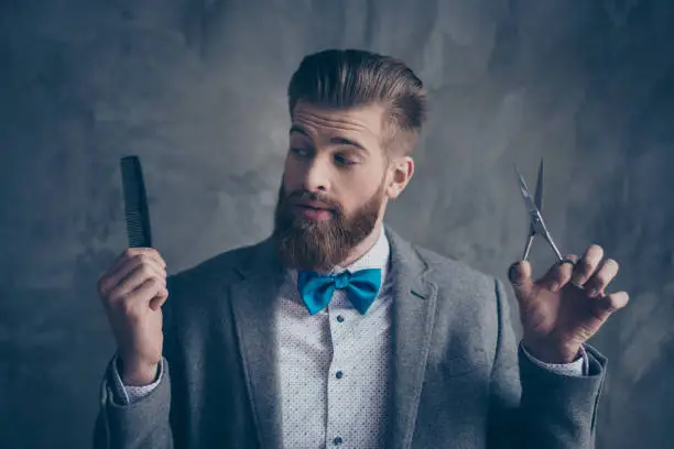 Portrait of Stylish young bearded man in a suit with bow-tie stands on a gray background and chooses between scissors and a comb. He solve going to the barbershop