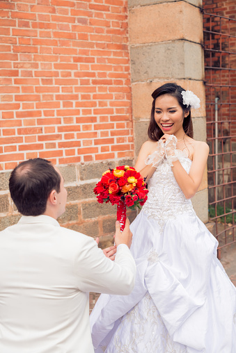 Back view of unrecognizable groom standing on one knee and holding colorful wedding bouquet in hand while his pretty Asian bride looking at him with toothy smile, brick outer wall on background
