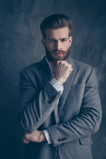 istock young stylish bearded guy with mustache in a suit stands on a gray background supporting his chin by fist 941792978