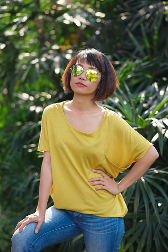 Confident young woman in sunglasses looking at camera while standing against bushy area illuminated with sunbeams, portrait shot