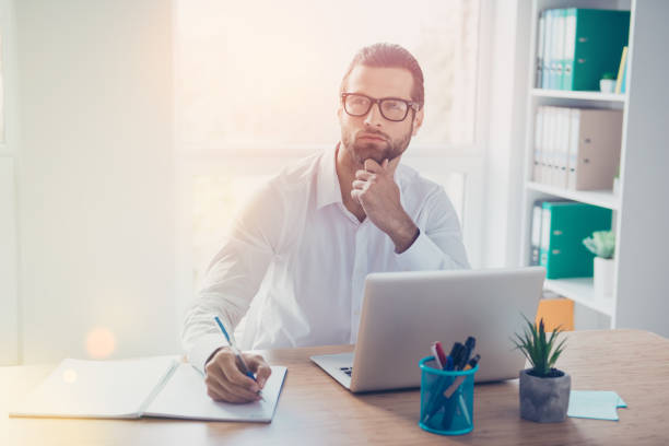 istock Handsome young businessman sits at the table in lightful office, dreaming and notes thought 941808408