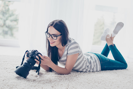 Young brunette pretty brunette photographer in spectacles checking photos on a camera while lying on the floor  at home