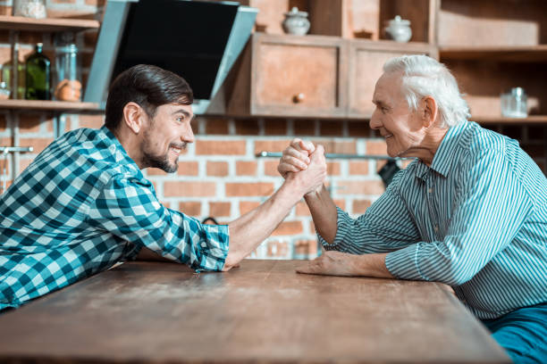 Nice happy man holding his fathers hand This is arm wrestling. Nice happy positive man holding his fathers hand and smiling while doing arm wrestling baby boomer stock pictures, royalty-free photos & images