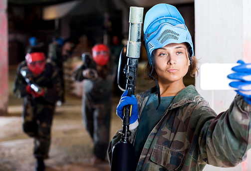 Young woman in blue mask is making selfie on battlefield of paintball club.