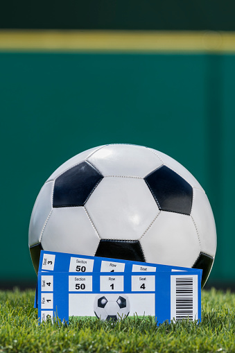 Two ticket stubs sitting in front of a black and white soccer ball that is lying in the grass of a stadium with a green padded wall in the background