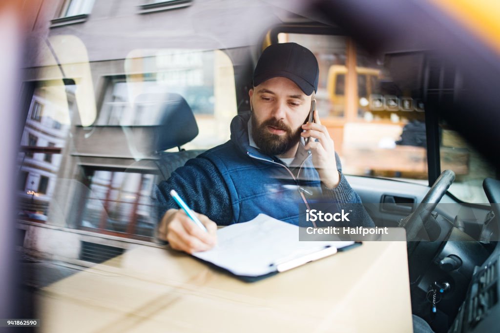 Delivery man with a parcel box in the car. Delivery man with a parcel box in the car- courier service concept. A man with a smartphone making a phone call. Shot through glass. Delivery Person Stock Photo