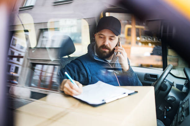 Delivery man with a parcel box in the car. Delivery man with a parcel box in the car- courier service concept. A man with a smartphone making a phone call. Shot through glass. messenger stock pictures, royalty-free photos & images